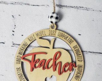 Può includere: Ciondolo in legno a forma di mela con la parola "Teacher" in lettere rosse. Il ciondolo è circondato da un cerchio con le parole "Mentor, Chaos Coordinator, Hard Working, Role Model, 2022 Supportive, Dedicated, Inspiring, Passionate".
