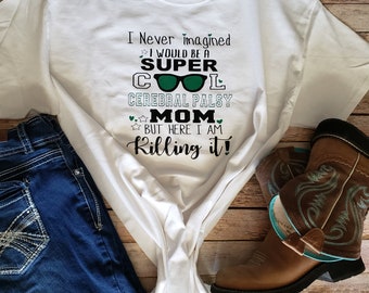 Op de afbeelding: Wit T-shirt met groene en zwarte tekst die luidt: "Ik had nooit gedacht dat ik een SUPER COOL Cerebral Palsy MOM zou zijn, maar hier ben ik en ik rock het!"