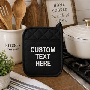 May include: A square black quilted pot holder featuring a pocket and the white text CUSTOM TEXT HERE printed in the center. The fabric has a diamond-stitched pattern and a small fabric loop at the top corner for hanging in a kitchen setting.