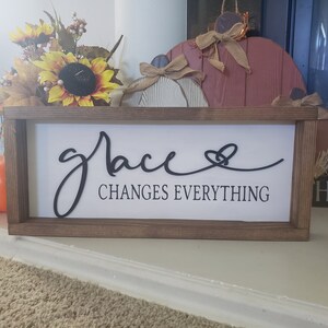 以下が含まれることがあります： 白い背景に黒い文字で「grace changes everything」と書かれた、木製のフレームの看板。ハートのマークも付いています。
