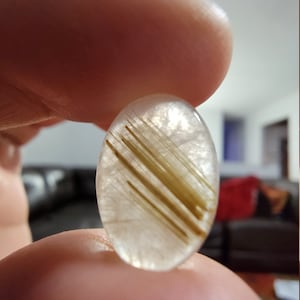 May include: A clear oval-shaped gemstone with thin, golden-brown inclusions. The gemstone is held between two fingers.