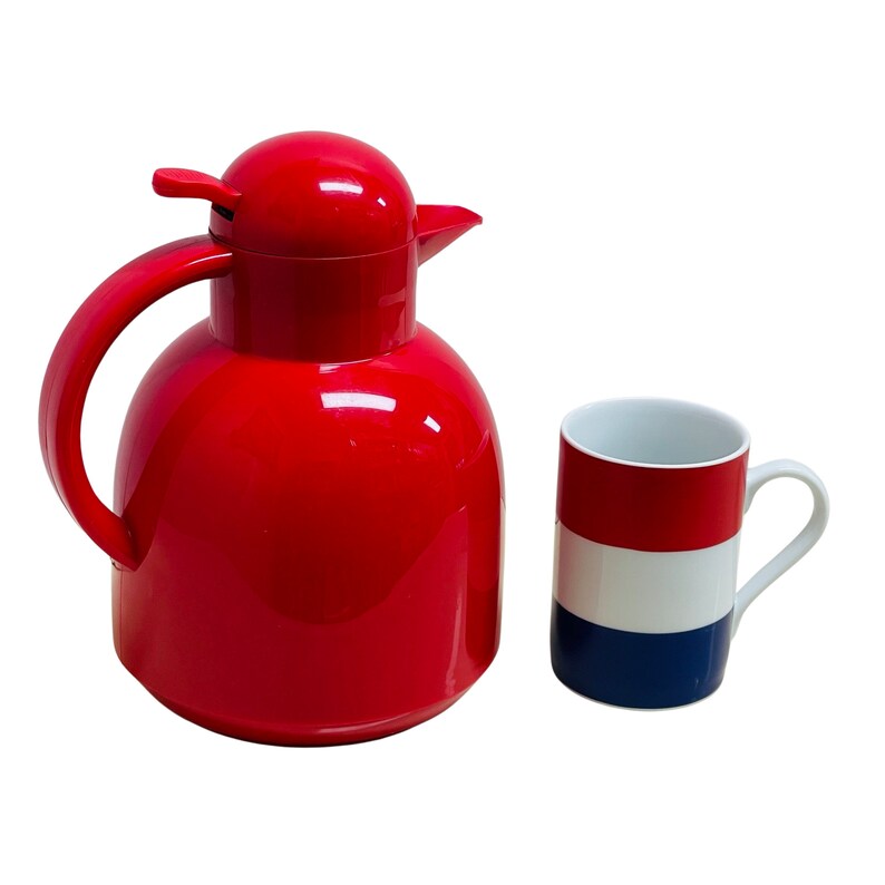 May include: A red thermal carafe with a lid and a handle, next to a white mug with red, white, and blue stripes.