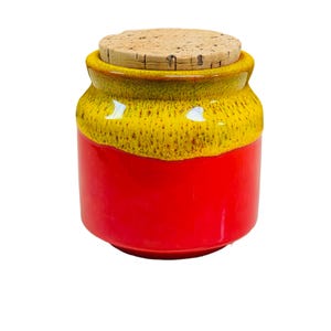 Könnte beinhalten: Eine rote und gelbe Keramikdose mit einem Korkdeckel. Die Dose hat eine gesprenkelte Glasur und eine abgerundete Form.