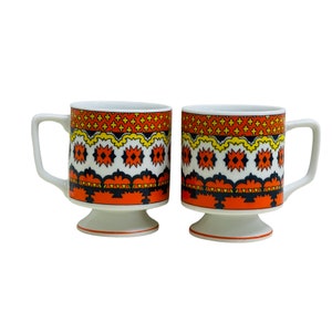 May include: Two white ceramic coffee mugs with a geometric pattern in orange, yellow, black, and white. The mugs have a pedestal base.