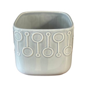 May include: A light blue ceramic planter with a square shape and a white geometric design. The planter has a smooth, glossy finish.
