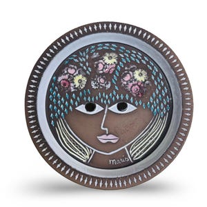 May include: A decorative ceramic plate with a stylized face, floral hair, and a diamond-patterned head covering. The plate has a brown background, white and brown border, and the name "Maris" is written on the bottom right.