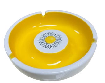 May include: A white ceramic ashtray with a yellow interior and a black and white daisy design in the center.