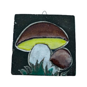 Op de afbeelding: Vierkante keramische tegel met een paddestoelontwerp. De paddenstoel heeft een bruine hoed, gele kieuwen en een witte steel, tegen een zwarte achtergrond. Groen gras bevindt zich aan de basis van de paddenstoel. Een draad is bevestigd om op te hangen.