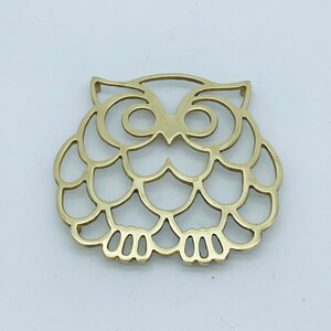Op de afbeelding: Gouden metalen onderzetter in de vorm van een uil met een uitgesneden ontwerp. De onderzetter heeft een gedetailleerd ontwerp van een uil met grote ogen en een gegolfd lichaam.