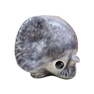 May include: A ceramic sculpture of a grey and white dinosaur head with a single large eye. The dinosaur has a textured surface and a small, pointed snout.