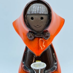 Op de afbeelding: Een keramiek beeldje van een persoon die een feloranje kapmantel en een bruine jurk draagt. De persoon heeft een bruin gezicht met witte ogen en een kleine glimlach. Het beeldje houdt een klein wit en bruin kopje vast.