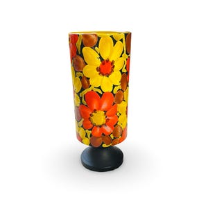 May include: A tall, cylindrical vase with a black base, adorned with a vibrant floral pattern. The flowers are in shades of yellow, orange, and brown against a black background. The vase is a decorative item.