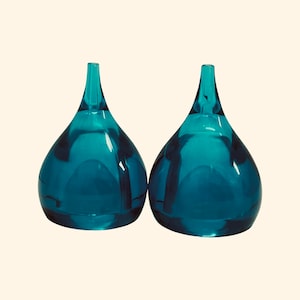 May include: Two teal glass vases, each with a teardrop-shaped body and a long, slender neck. The vases are set against a light background, accentuating their smooth, reflective surfaces. The design is modern and elegant.