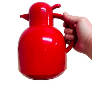 May include: A bright red thermos with a bird-shaped handle and lid. The thermos is made of plastic and has a glossy finish.