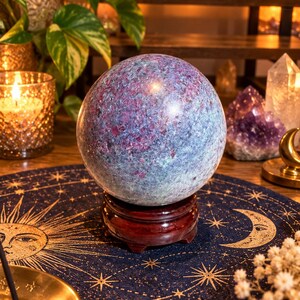 May include: A polished, spherical ruby in fuchsite crystal resting on a dark, circular wooden stand. The stone features a mottled texture with deep magenta and red ruby inclusions embedded within a cool, blue-green fuchsite matrix.
