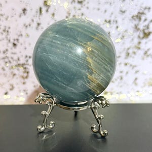 May include: A blue-green crystal sphere with gold-colored streaks, displayed on a decorative silver-colored metal stand. The sphere has a polished surface and is a focal point against a blurred background.