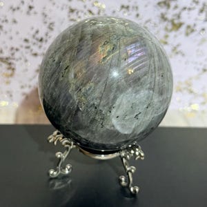 May include: A gray labradorite sphere with iridescent flashes of color, displayed on a decorative silver-toned metal stand. The sphere has a polished surface and is a natural stone. The stand has a detailed, ornate design.