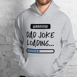 May include: A light gray hoodie with a front pocket and a drawstring hood. The hoodie features the text "WARNING! DAD JOKE LOADING..." in black, with a blue loading bar graphic. The person in the image is smiling.