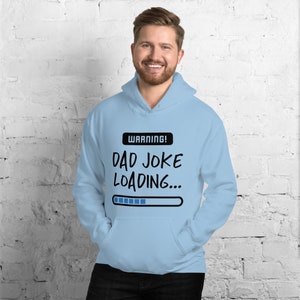 May include: Light blue hoodie with a front pocket and a drawstring hood. The hoodie features the text "WARNING! DAD JOKE LOADING..." above a loading bar graphic in black and white. The person in the image is smiling.