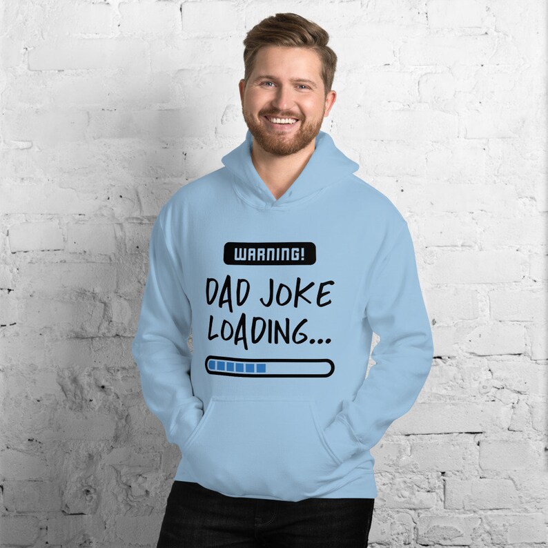 May include: Light blue hoodie with a front pocket and a drawstring hood. The hoodie features the text "WARNING! DAD JOKE LOADING..." above a loading bar graphic in black and white. The person in the image is smiling.