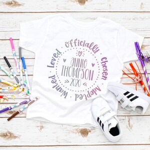 Puede incluir: Camiseta blanca con un dise&ntilde;o circular morado y rosa que dice "Oficialmente Elegido, Deseado, Amado, Adoptado, Annie Thompson, 2020".