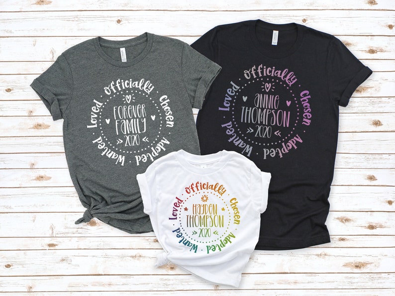 Puede incluir: Tres camisetas con un dise&ntilde;o circular que dice "Loved Officially Chosen Adopted Wanted" con el a&ntilde;o 2020 y un nombre en el centro. La primera camiseta es gris con texto blanco, la segunda es negra con texto rosa y la tercera es blanca con texto arco&iacute;ris. Los nombres en las camisetas son "Forever Family", "Annie Thompson" y "Hayden Thompson".