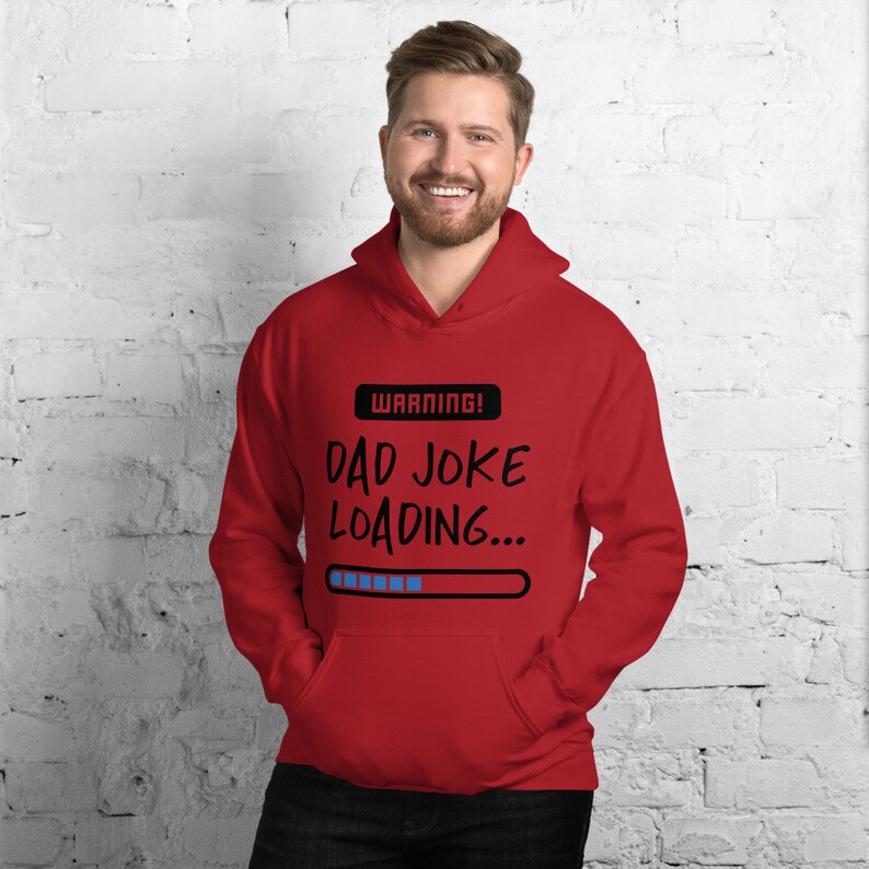 May include: Red hooded sweatshirt with the text "WARNING! DAD JOKE LOADING..." in black. The design includes a loading bar graphic. The sweatshirt has a front pocket and a drawstring hood.