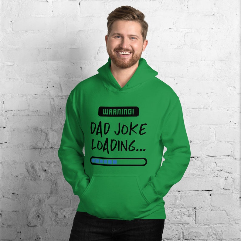 May include: A green hooded sweatshirt with the text "WARNING! DAD JOKE LOADING..." in black and white. The sweatshirt has a front pocket and a drawstring hood. The person in the image is smiling.