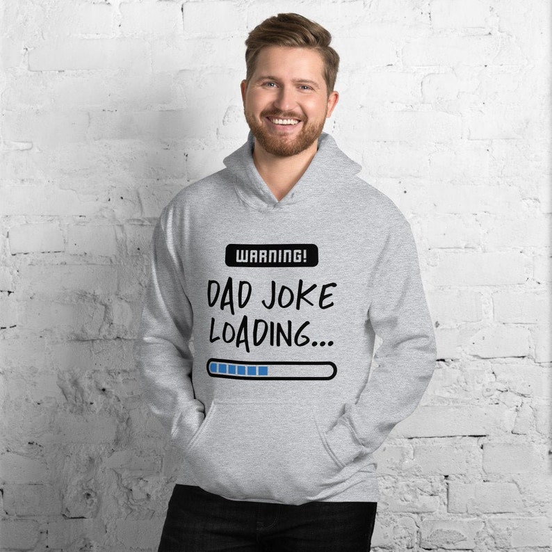May include: A light gray hoodie with a front pocket and a drawstring hood. The hoodie features the text "WARNING! DAD JOKE LOADING..." in black, with a blue loading bar graphic. The person in the image is smiling.