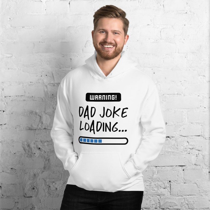 May include: White hoodie with a front pocket and a drawstring hood. The hoodie features the text "WARNING! DAD JOKE LOADING..." in black, with a blue progress bar graphic. The person in the image is smiling.