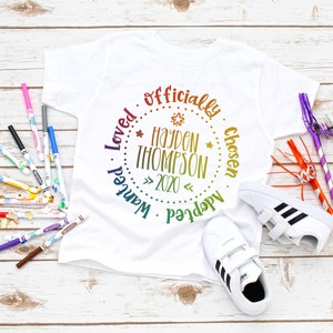 Puede incluir: Camiseta blanca con un dise&ntilde;o circular de arco&iacute;ris que dice "Adoptado oficialmente, elegido, amado, querido" con el nombre "Hayden Thompson" y el a&ntilde;o "2020" en el centro.