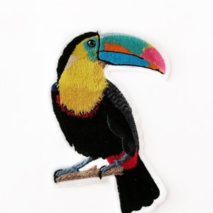 Op de afbeelding: Geborduurde strijk-patch van een toekan die op een tak zit. De toekan heeft een zwart lichaam, een gele borst, een rode keel en een kleurrijke snavel met blauwe, oranje en roze accenten.