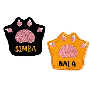 Puede incluir: Dos parches con forma de huella de pata, uno negro con el nombre "SIMBA" y otro naranja con el nombre "NALA".