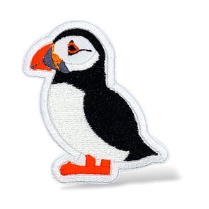 May include: Embroidered patch featuring a puffin bird. The patch has a white outline, with the bird's body in black and white. The beak is orange, and the feet are orange. The patch is designed to be ironed or sewn onto fabric.