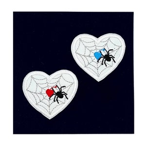 May include: Two white felt heart-shaped patches with embroidered spiderwebs and black spiders. One spider has a red heart in its web, and the other has a blue heart.