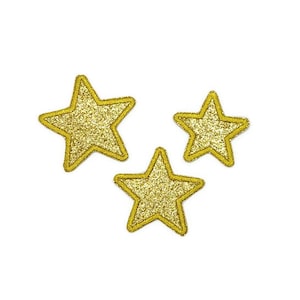 Puede incluir: Tres estrellas doradas con purpurina y contornos amarillos. Las estrellas están dispuestas en un grupo, con la estrella más grande en la parte superior y las dos estrellas más pequeñas debajo.