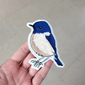 Puede incluir: Un parche bordado de un pájaro azul y marrón con costuras blancas alrededor de los bordes.