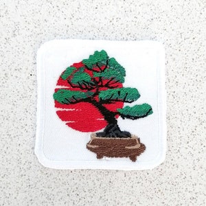 May include: Embroidered patch featuring a green bonsai tree in a brown pot, with a red sun rising behind it. The patch is white with a white border.