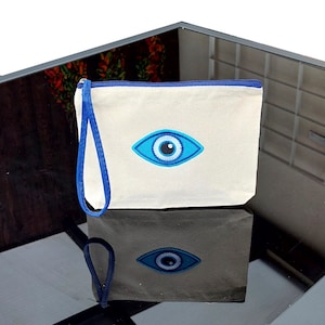 Puede incluir: Una bolsa de lona blanca con cremallera con un diseño de ojo azul malvado. La bolsa tiene una correa de tela azul.
