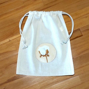 May include: A white cotton drawstring bag with a round embroidered design of a white cotton boll on a brown stem.