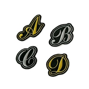 Op de afbeelding: Vier strijkbare patches met de letters A, B, C en D in een cursief lettertype. De letters zijn zwart omrand en gevuld met gouden of zilveren draad.