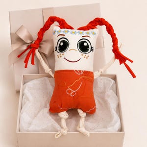 Può includere: Bambola di peluche fatta a mano con viso bianco, occhi grandi e capelli rossi intrecciati. La bambola indossa un completo arancione ed è presentata in una confezione regalo con un fiocco. Le braccia e le gambe della bambola sono in tessuto color crema.