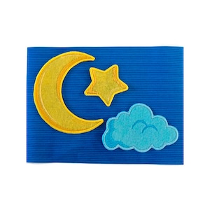 Pode incluir: Um fundo de feltro azul com uma lua crescente amarela, uma estrela amarela e uma nuvem azul. A lua e a estrela são bordadas.