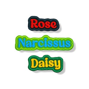 Puede incluir: Tres parches bordados con nombres de flores. El parche superior dice "Rose" en letras rojas con un borde negro y azul. El parche del medio dice "Narcissus" en letras azules con un borde verde. El parche inferior dice "Daisy" en letras amarillas con un borde verde.