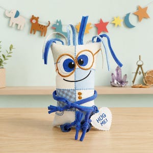 Può includere: Un giocattolo di peluche stravagante in stile patchwork in crema, blu e beige. Presenta grandi occhi blu, occhiali e un viso sorridente. Il giocattolo ha accenti blu, tra cui capelli a cappio e una cintura annodata. Un'etichetta a forma di cuore recita "HUG ME!"