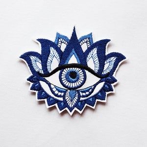 May include: Embroidered patch featuring a blue and white eye design, surrounded by a stylized lotus flower. The eye has a black eyebrow and a blue iris. The patch is on a white background.