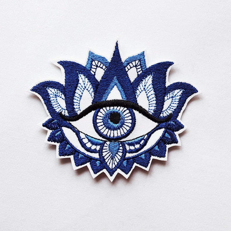 May include: Embroidered patch featuring a blue and white eye design, surrounded by a stylized lotus flower. The eye has a black eyebrow and a blue iris. The patch is on a white background.