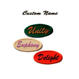 Puede incluir: Tres parches bordados de forma ovalada con diferentes nombres. El parche superior es verde con la palabra "Unity" en naranja. El parche del medio es beige con la palabra "Euphony" en rosa. El parche inferior es rojo con la palabra "Delight" en negro.