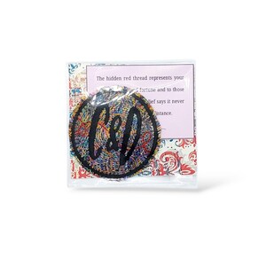 Pode incluir: Um patch circular colorido com as letras "C&D" em escrita preta, embalado em uma manga de pl&aacute;stico transparente. O patch &eacute; feito de fios entrela&ccedil;ados em v&aacute;rias cores. Um cart&atilde;o rosa com texto &eacute; vis&iacute;vel atr&aacute;s do patch.