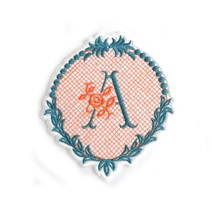 Può includere: Una toppa ricamata in blu turchese con sfondo bianco e un motivo floreale color pesca. La toppa presenta la lettera "A" in un carattere serif.